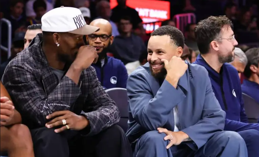 The Golden State Warriors players Jimmy Butler and Stephen Curry sitting courtside.